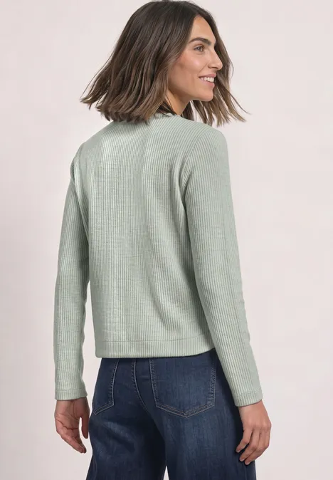 Vrouw van achteren in lichtgroene trui en blauwe jeans, ze draait het hoofd en glimlacht zacht.