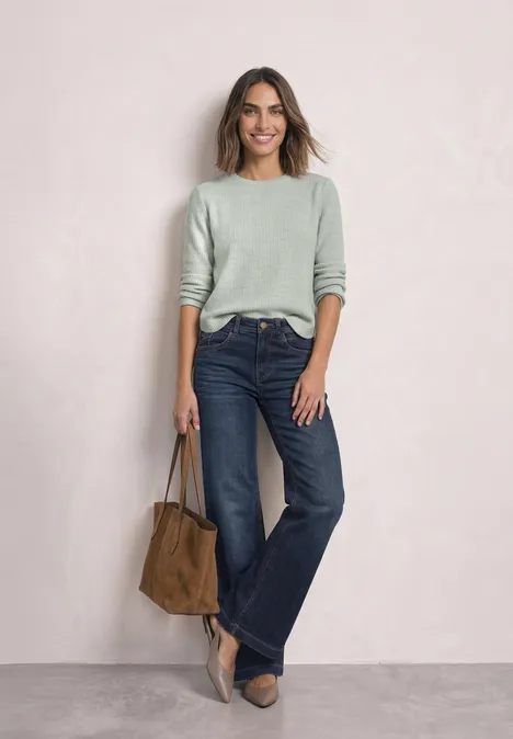 Een vrouw in een groen trui, wijde jeans, hakken en suède tas voor een neutrale achtergrond.