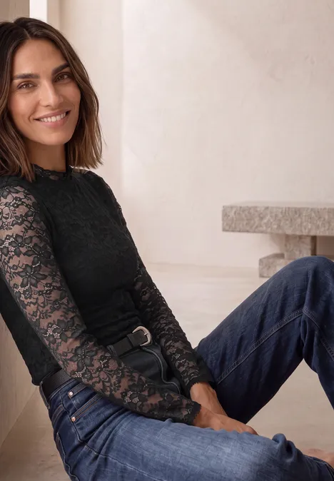 Eine lächelnde Frau in schwarzer Spitzenbluse und Jeans sitzt entspannt vor einer hellen Wand.
