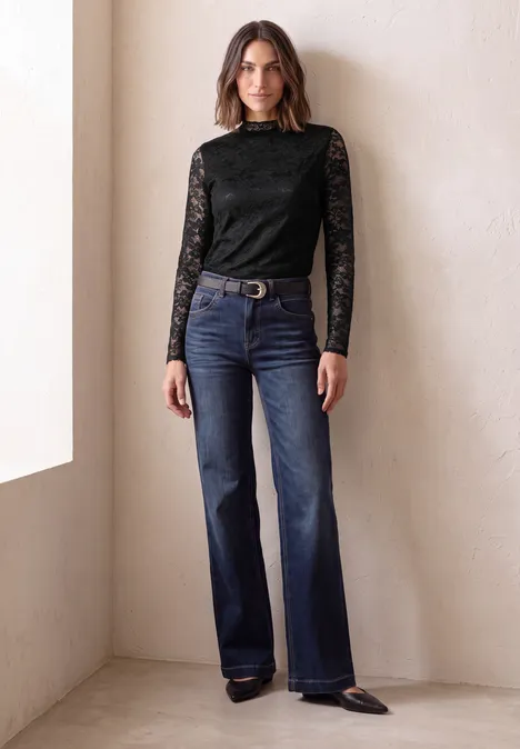 Frau in schwarzer Spitzenbluse und weitem Jeans steht vor heller Wand.