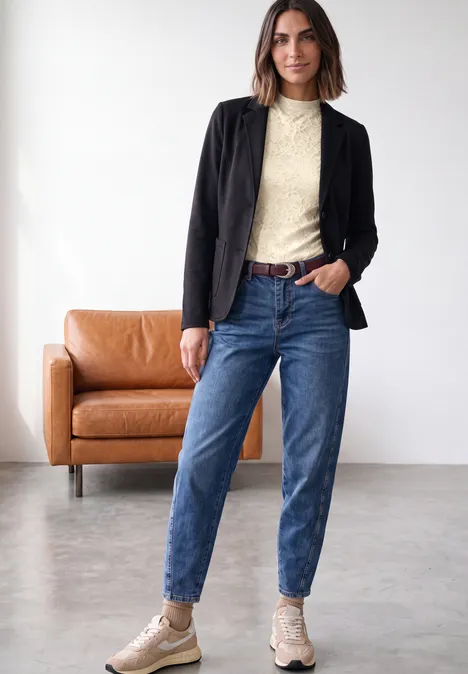 Eine Frau in schwarzer Jacke, cremefarbenem Pullover, blauen Jeans und beigen Boots steht in modernem Raum.
