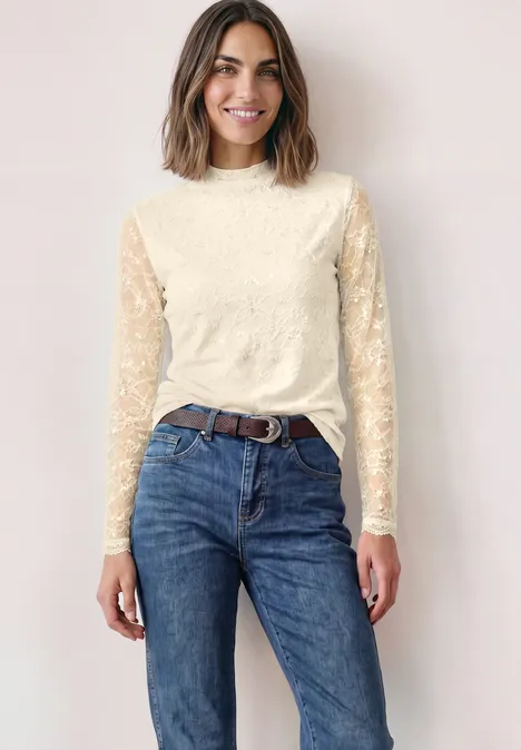 Frau mit braunen Haaren trägt cremefarbenes Spitzenoberteil und blaue Jeans, lächelnd vor hellem Hintergrund.