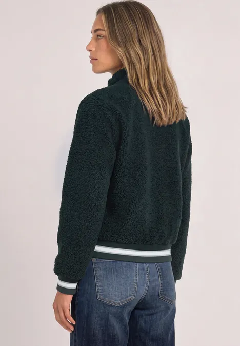 Frau von hinten trägt eine schwarze Teddyjacke und Jeans, schaut seitlich vor hellem Hintergrund.