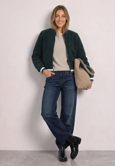 Frau lehnt an Wand, trägt Bomberjacke, Pulli, Jeans, Stiefel und hält Tasche.