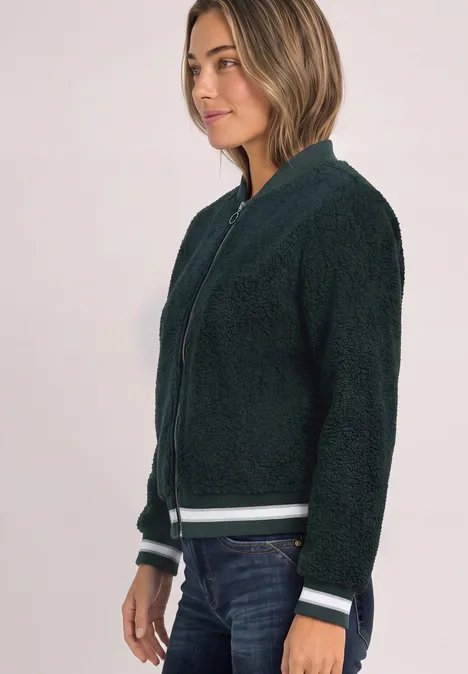 Eine Frau in dunklem Teddy-Blouson mit weiß-grünen Streifen am Bund, lässig zur Jeans.