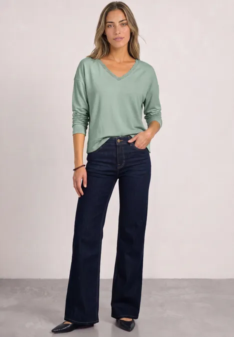 Een vrouw in lichtgroene trui en donkerblauwe uitlopende jeans staat nonchalant.