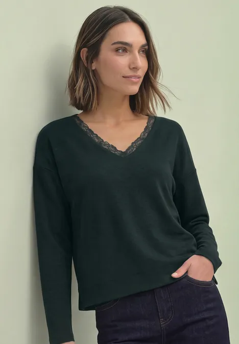 Frau in schwarzem Langarmshirt mit Spitzen-V-Ausschnitt und Jeans, steht entspannt mit Hand in Tasche.