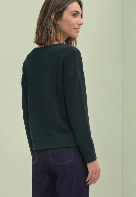 Frau in dunkelgrünem Pullover und Jeans, von hinten vor hellem Hintergrund.