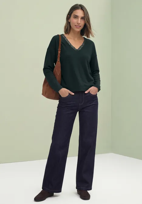Frau in grünem Pullover, weiten Jeans und brauner Tasche vor hellgrünem Hintergrund.