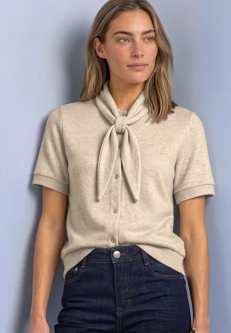 Kurzarm Jacke mit Schalkragen clay sand mel. Frau in beiger Kurzarm-Strickbluse mit Schleifenkragen, dunkelblaue Jeans, vor hellblauem Hintergrund.