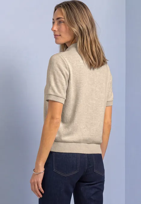 Kurzarm Jacke mit Schalkragen clay sand mel. Frau in beigeem Kurzarm-Pullover und blauen Jeans im seitlichen Profil vor hellem Hintergrund.