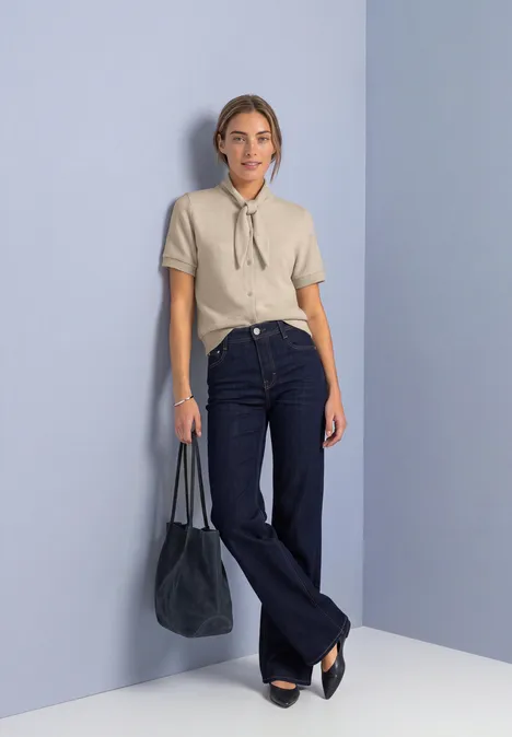 Kurzarm Jacke mit Schalkragen clay sand mel. Frau in beiger Bluse mit Schluppe, dunkelblaue Schlagjeans und schwarzer Tasche vor pastellfarbener Wand.