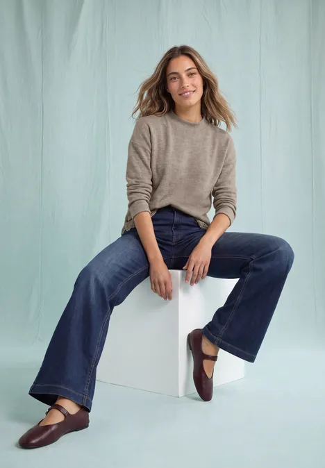 Een lachende vrouw zit op een wit kubusje, draagt een grijze trui, wijde jeans en bruine ballerina’s.
