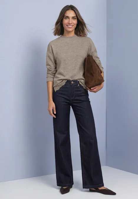 Vrouw in beige trui en donkere wijde jeans met bruine tas voor effen achtergrond.