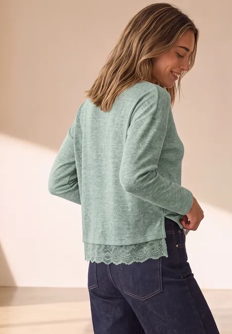 Vrouw in profiel met groene trui met kanten zoom en spijkerbroek, glimlachend.