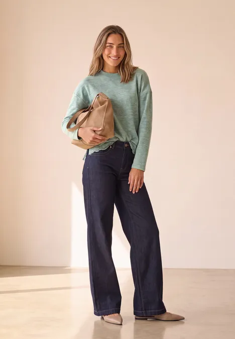 Een glimlachende vrouw draagt een mintgroene trui, wijde jeans en houdt een bruine tas in een lichte kamer.