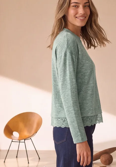 Een vrouw op de foto draagt een groen shirt met lange mouwen en kanten zoom en een donkere broek.