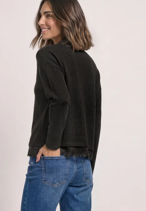 Vrouw in zwart langarmshirt en blauwe spijkerbroek, glimlachend zijwaarts kijkend.