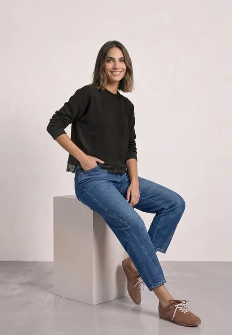 Vrouw zittend op een wit blok, draagt zwarte trui, blauwe jeans en bruine schoenen, lichte glimlach.