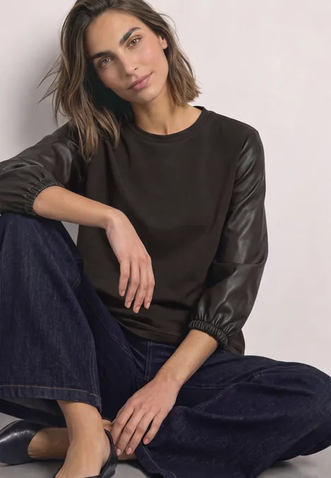 Shirt mit 3/4-Ärmeln in Leder-Optik black coffee Frau sitzt lässig, trägt braunen Pullover mit Lederärmeln, weite Jeanshose und Sandalen vor heller Wand.