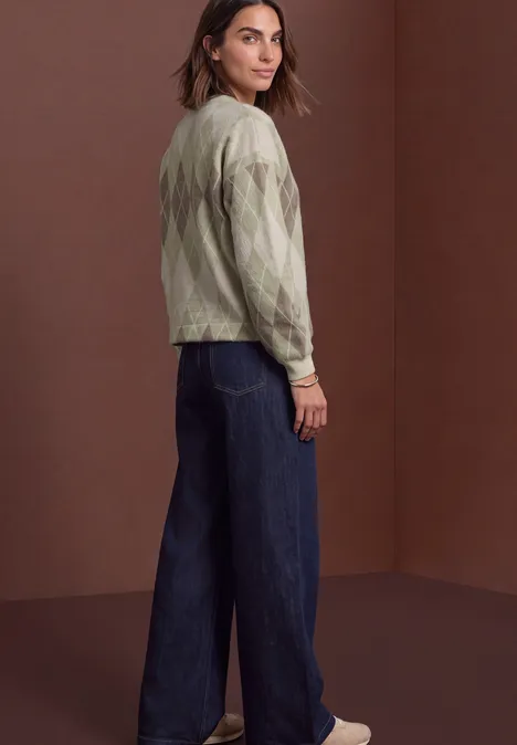 Shirt met lange mouwen, ronde hals en ruitjespatroon eucalyptus green Vrouw in groen, gemêleerde trui, wijde jeans en witte sneakers, staand voor een bruine achtergrond en achteromkijkend.