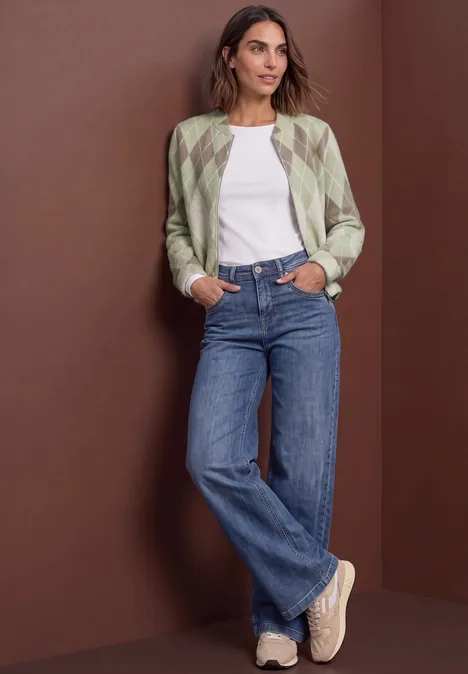 Blouson met lange mouwen en ruitjespatroon eucalyptus green Vrouw leunt tegen bruin muurtje, met gemêleerd jack, wit T-shirt, losse jeans en comfortabele sneakers.