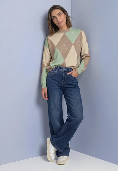 Langarm Shirt mit Turtle Neck und Muster eucalyptus green Frau in pastellfarbenem Argyle-Pullover, weiten Jeans und weißen Sneakern vor schlichter Wand.