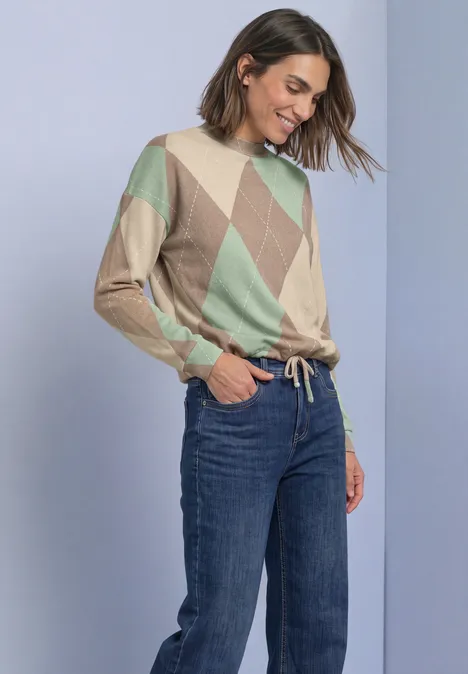 Langarm Shirt mit Turtle Neck und Muster eucalyptus green Frau in Jeans und Pullover mit Rautenmuster, lächelt, Hände in den Hosentaschen vor neutralem Hintergrund.
