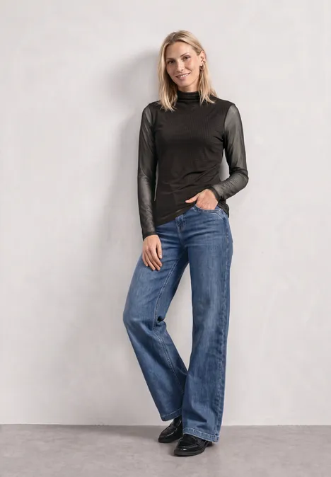 Frau vor neutralem Hintergrund trägt schwarzen Langarmtop, weite blaue Jeans und schwarze Schuhe.