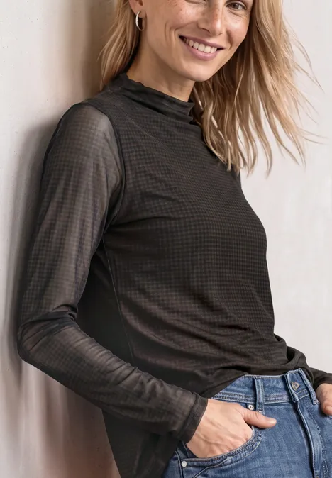 Frau lehnt an Wand, trägt schwarzen transparenten Langarmpullover und Jeans, lächelt leicht.