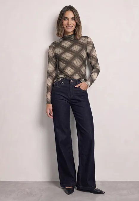 Vrouw in geruit coltrui en wijde, donkere broek, glimlachend voor een lichte achtergrond.