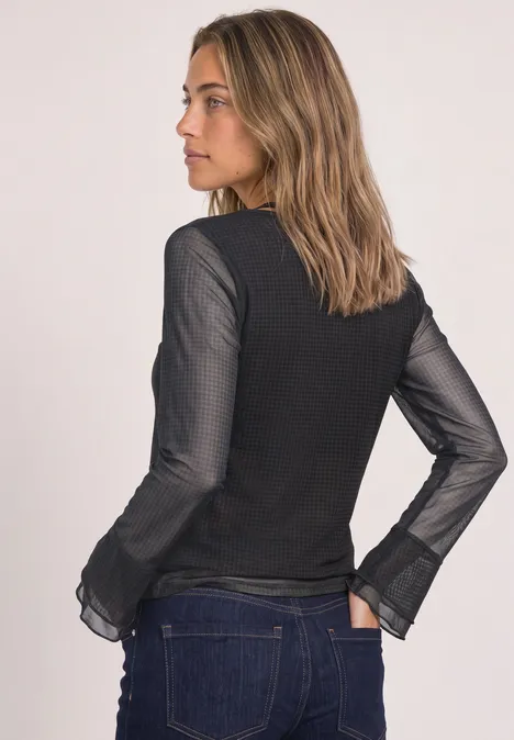 Eine Frau steht seitlich, trägt einen schwarzen Pullover mit transparenten Ärmeln und blaue Jeans.