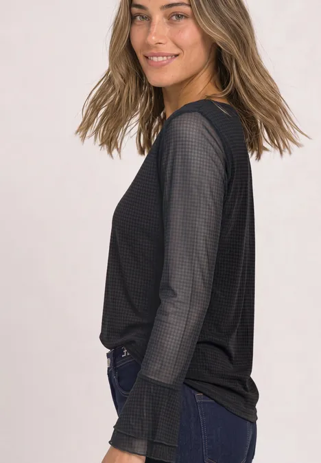 Junge Frau im schwarzen Mesh-Oberteil und Jeans, seitlich stehend und lächelnd.