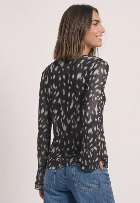 Frau in schwarzer, gemusterter Bluse und Jeans, seitlich fotografiert.