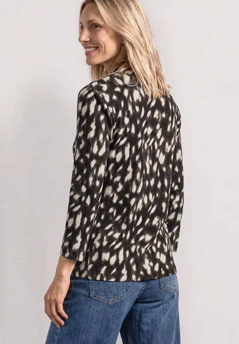 Frau in schwarz-weiß gemusterter Bluse und Jeans schaut lächelnd über die Schulter.