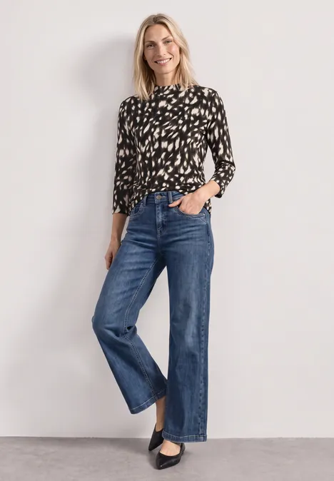 Frau mit schwarzer, gemusterter Bluse, weiter blauer Jeans und schwarzen Pumps vor hellem Hintergrund.