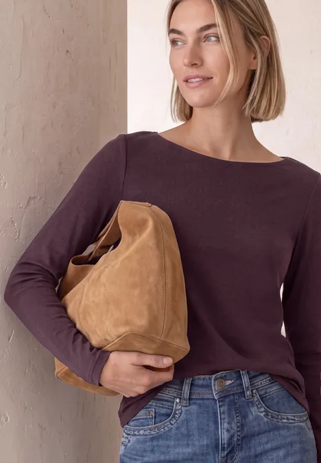 Frau in dunkelrotem Pullover und Jeans trägt eine große, braune Wildledertasche.