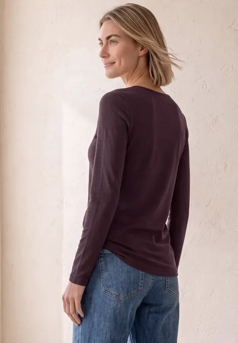 Frau von hinten/seitlich in dunkelviolettem Langarmshirt und blauen Jeans vor hellem Hintergrund.