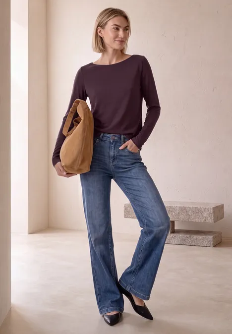 Frau in lila Langarmshirt, blauen Jeans und schwarzen Ballerinas, hält eine braune Tasche.