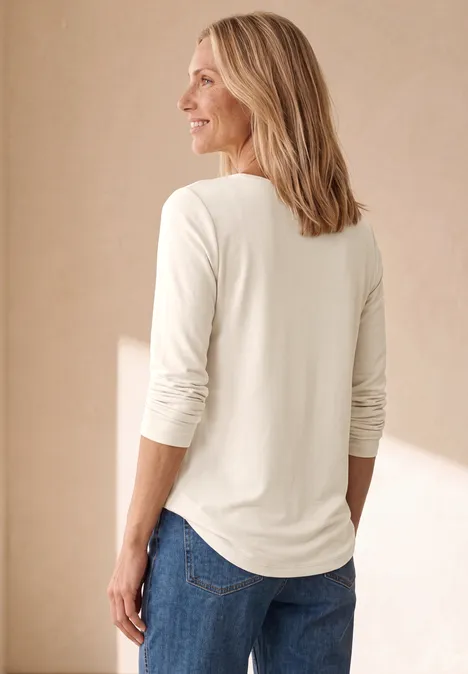 Eine Frau in weißem Langarmshirt und blauer Jeans steht seitlich vor hellem Hintergrund.