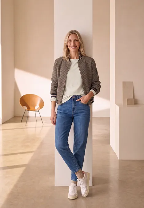 Eine lächelnde Frau in Jeans und Sneakern lehnt in einem hellen, modernen Raum an einer Säule.