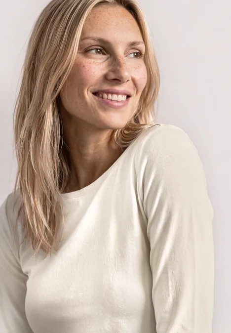 Eine blondhaarige Frau in weißem Shirt lächelt seitlich vor hellem Hintergrund.