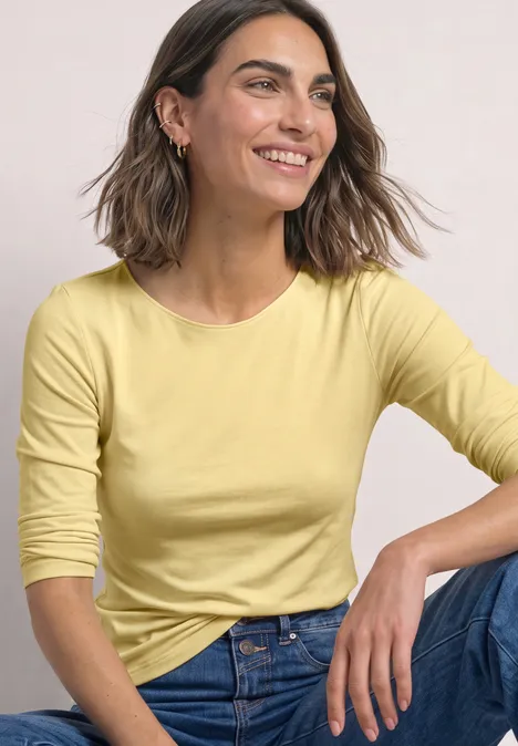 Basic Shirt mit U-Boot-Ausschnitt sunbeam yellow Frau in gelbem Oberteil und Jeans, sitzt lächelnd.