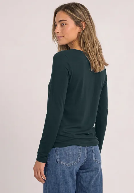 Junge Frau im Profil mit dunkelgrünem Langarmshirt und blauen Jeans vor hellem Hintergrund.