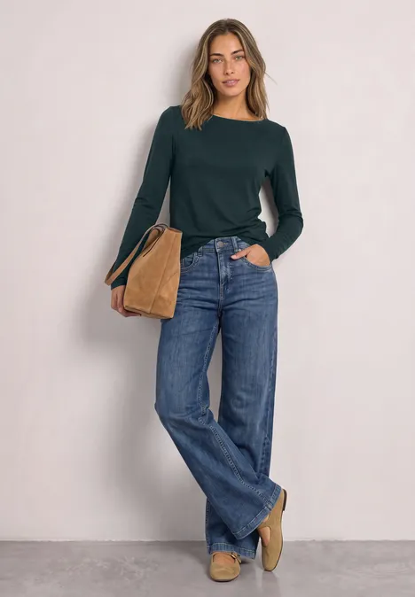 Frau in dunkelgrünem Pullover, weiten Jeans und beiger Tasche vor neutraler Wand.