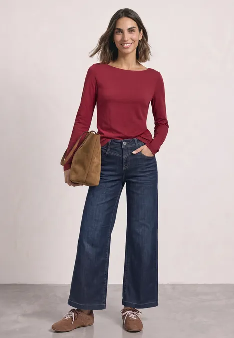 Frau in rotem Langarmshirt mit weiten Jeans, braunen Schuhen und Tasche vor hellem Hintergrund.