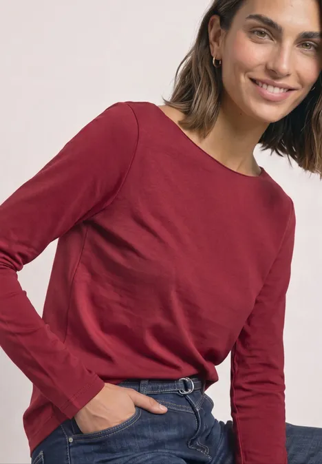 Lächelnde Frau in rotem Langarmshirt und Jeans, eine Hand in der Tasche.