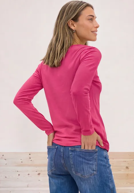 Frau von hinten in pinkem Langarmshirt und Jeans, Hände in den Taschen, vor hellem Hintergrund.