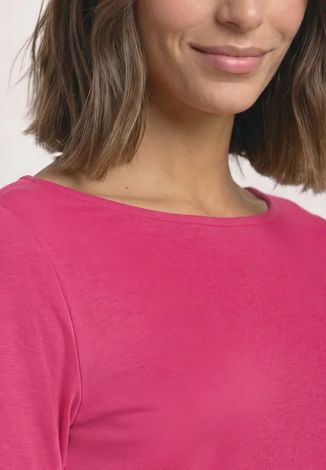Eine Frau mit schulterlangem Haar trägt ein pinkes Langarmshirt mit Boot-Ausschnitt und dezentem Lächeln.