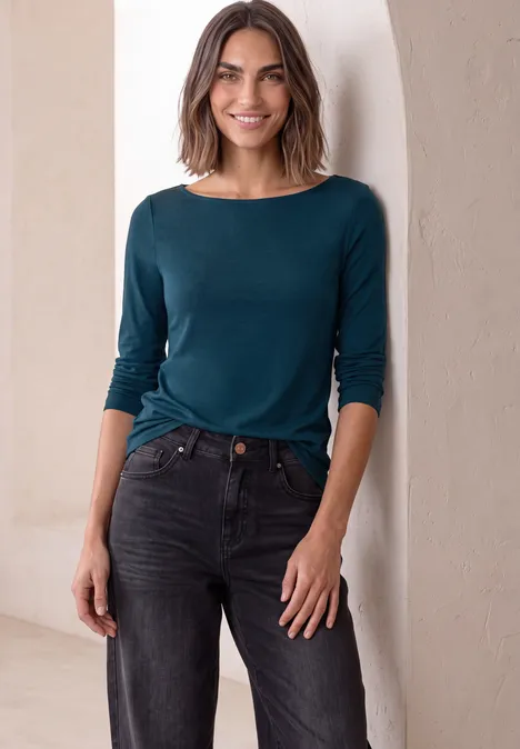 Frau mit schulterlangem Haar trägt petrolfarbenes Shirt und schwarze Jeans, lächelnd in einem hellen Raum.